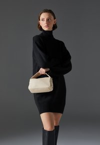 Black oversized sweater dress, smooth beige handbag with structured shape and a curved handle, worn with knee-high black boots.