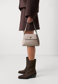 Borsa beige con un design strutturato, rifiniture nere e hardware in argento. Indossata con un cappotto marrone e stivaletti coordinati con cuciture decorative.