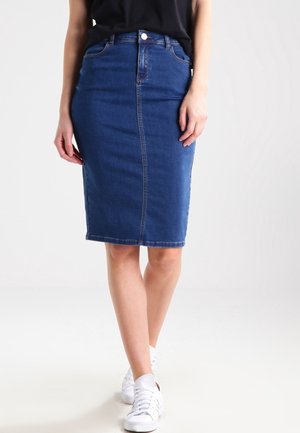 Femme portant une jupe crayon en denim bleu jusqu'aux genoux, associée à des baskets blanches et un haut noir, debout devant un fond uni.