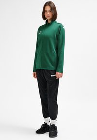 Grön zip-up jacka med hög krage, kombinerad med svarta träningsbyxor med en vit logotyp. Svarta skor och vita strumpor fullbordar outfiten.