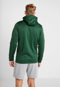 Homme portant un sweat à capuche vert foncé et un short gris clair, debout de dos sur un fond clair uni.