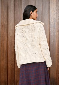 Vastag krémszínű, kábelmintás kötött kardigán széles bordás gallérral, texturált mintákkal az ujjakon és a testén, kockás szoknyával párosítva.