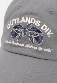 Căciulă de baseball din bumbac gri, cu text brodat "OUTLANDS DIV." și design floral în nuanțe de bleumarin. Are o vizieră curbată și o suprafață texturată.
