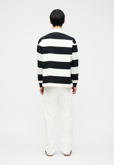 Άνδρας που έχει την πλάτη γυρισμένη, φοράει μαύρο και λευκό οριζόντια ριγέ πουκάμισο με μακριά μανίκια και άσπρο παντελόνι, πάνω σε απλό λευκό φόντο.