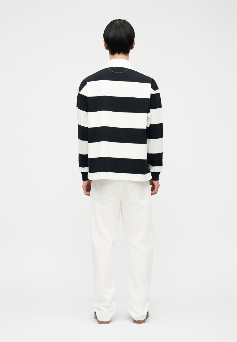 Homme de dos portant un haut à manches longues rayé horizontalement noir et blanc et un pantalon blanc sur un fond blanc uni.