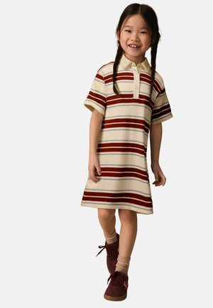 Junges Mädchen mit geflochtenem Haar, das ein kurzärmeliges, gestreiftes Polo-Kleid und bordeauxfarbene Sneaker trägt, lächelt und geht nach vorne.