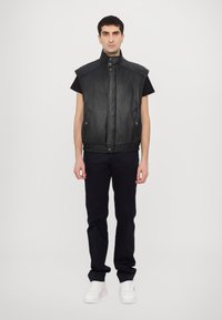 Μαύρο δερμάτινο γιλέκο χωρίς μανίκια με ψηλό γιακά, μπροστινό φερμουάρ και κουμπωτές κλείσεις; φορεμένο πάνω από μαύρο πουκάμισο και συνδυασμένο με σκούρα παντελόνια και λευκά αθλητικά παπούτσια.