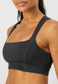 Sostenedor deportivo negro fabricado con tejido elástico, presenta un escote cuadrado, tirantes gruesos y detalles en las costuras que aportan estructura adicional.