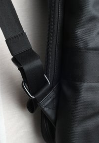Schwarze Tasche aus strukturiertem Stoff, mit einem Reißverschluss, verstellbarem Schultergurt und metallicfarbenen Beschlägen. Nahaufnahme, die sich auf die Details des Gurtes konzentriert.