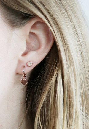 Roze gouden oorbellen met een ronde stud en een hangend hartje, beide met een gepolijste afwerking, gedragen in het oor van een model.