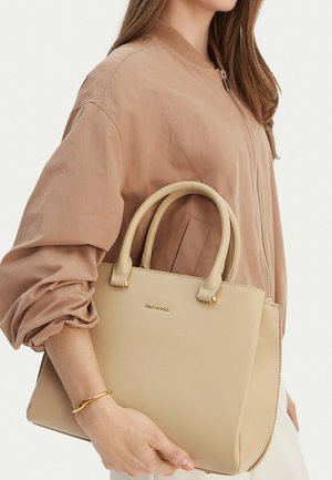 Frau in beiger Jacke hält eine cremefarbene Gino Rossi Handtasche mit doppelten Griffen und goldenem Logo, trägt ein wellenförmiges goldenes Armband.