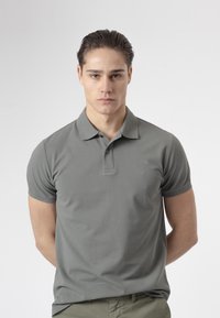 Joven con el pelo peinado hacia atrás, vistiendo una camisa polo gris y pantalones oliva, de pie con las manos detrás de la espalda against a fondo liso.