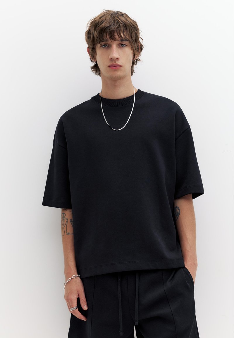 Jeune personne aux cheveux mi-longs portant un t-shirt noir ample et un pantalon noir, accessoirisée d'un collier et d'un bracelet en chaîne argentés.