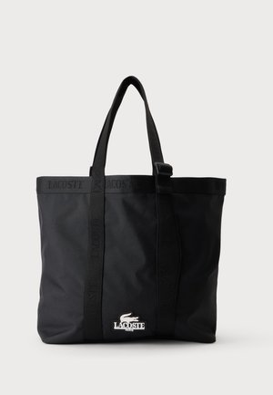 Czarna torba typu tote Lacoste z podwójnymi uchwytami, markowymi paskami oraz białą naszywką z logo krokodyla na przedniej dolnej części na środku.