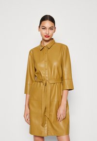 Abito camicia in pelle color senape con colletto, chiusura a bottoni, maniche a tre quarti e cintura annodata in vita. Tessuto liscio con cuciture.