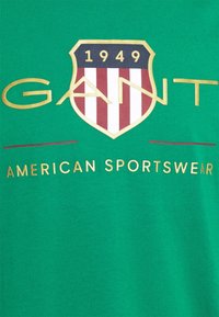 Zielona bawełniana koszulka z dużym logo w kształcie tarczy, z czerwonymi i białymi paskami, złotym napisem "GANT" oraz "AMERICAN SPORTSWEAR" oraz rokiem "1949".