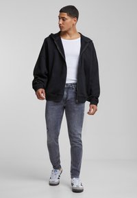 Crna zip-up dukserica sa opuštenim fitom, kombinirana sa sivim iznošenim trapericama i bijelim tenisicama s crnim detaljima, na neutralnoj pozadini.