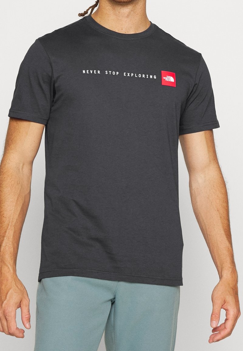 Hombre con camiseta de manga corta gris oscuro con el texto blanco "Never Stop Exploring" y el logo rojo de The North Face en el pecho.