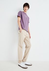 Purple cotton t-shirt with front logo, beige cotton trousers, and white sneakers with gray accents, standing against a plain white background.