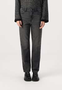 Dunkelgraue Jeans aus Denim mit geradem Schnitt, fünf Taschen und dezentem Fade. Kombiniert mit schwarzen Schuhen und einem gemusterten Pullover.