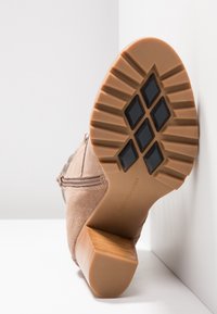 Chaussure en daim marron avec une semelle en caoutchouc, présentant un dessous texturé et un talon en bois. Fermeture éclair sur le côté avec des accents décoratifs.