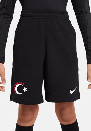 Dziecko w czarnych spodenkach Nike z białym logo swoosh i półksiężycem z gwiazdą, „AY-YILDIZLILAR” na czerwono, stoi wyprostowane.