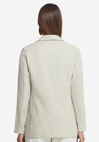 Beige kavaj med en strukturerad textil med små, mönstrade detaljer, notch-revers och långa ärmar. Bakstycket är figursytt med en enda ventil.