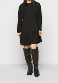 Černé dlouhé šaty s vrstveným lemem, vyrobené z texturovaného materiálu, spojené s vysokými černými koženými botami.