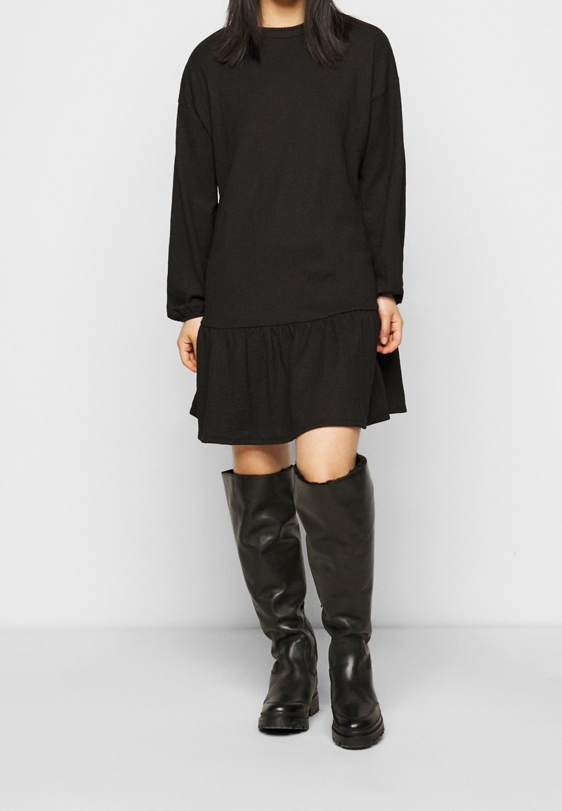 Černé dlouhé šaty s vrstveným lemem, vyrobené z texturovaného materiálu, spojené s vysokými černými koženými botami.