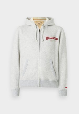Gaiši pelēks zip-up hoody ar priekšējiem kabatām, kapuci ar auklām, sarkano "Hollister California" logo uz krūtīm un mazu sarkano putnu logo uz piedurknes cuff.