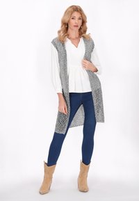 Cardigan lung gri tricotat cu glugă, purtat peste un top alb peplum, combinat cu blugi albastri și cizme de cowboy bej. Textură netedă, aspect stratificat.