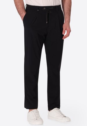 Uomo che indossa pantaloni neri su misura con vita con bottone e coulisse, abbinati a sneaker bianche e una camicia bianca infilata dentro.