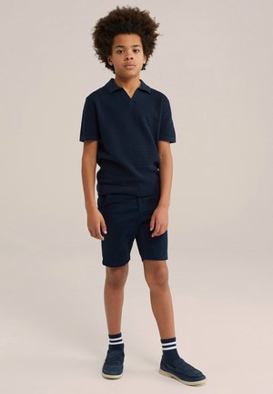 Polo en maille bleu marine avec un col en V, associé à un short bleu marine. Porté avec des chaussettes rayées bleu marine et blanches et des chaussures décontractées bleu foncé.