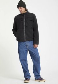 Zwarte fleecejas met ritssluiting aan de voorkant, twee borstzakken en een hoge kraag, gecombineerd met losse blauwe spijkerbroek en donkere sneakers.