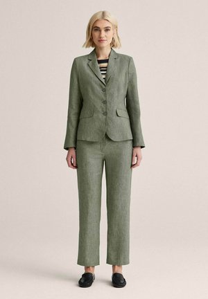 Kvinde iført olivengrøn skræddersyet jakkesæt med tredobbelt knapblazer og bukser med lige ben, kombineret med sorte slip-on sko og en stribet top.