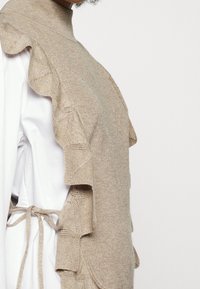 Beige neulepaita, jossa on korkeakaulus ja röyhelöä sivuilla. Siinä on teksturoitu pinta ja nauhat takana.