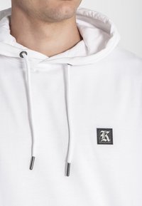 Sudadera blanca con capucha ajustable, que presenta un pequeño parche negro cuadrado con un logo blanco de "K" y puntas de metal plateadas en los cordones.