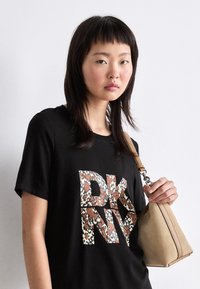 Černé tričko s velkým texturovaným logem "DKNY" v designu mnohobarevného leopardího potisku. Modelka drží béžovou kabelku.