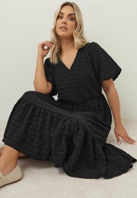 Musta, kuvioitu maksimekko, jossa on V-kaula-aukko, lyhyet hihat ja gathered-hameominaisuus. Kangas on ryppyistä ja tarjoaa liikettä sekä volyymia.