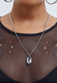 Colar de corrente prateada com um pendente retangular apresentando um design de silhueta preta, repousando sobre um top de malha preta transparente com acentos coloridos.