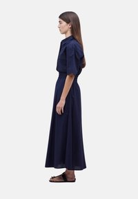 Vestido maxi azul marino con mangas cortas abullonadas, cierre frontal con botones y cintura elástica, confeccionado en una tela texturizada. Se presentan sandalias planas.