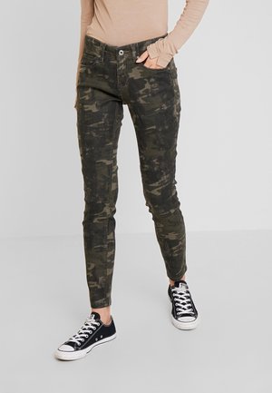 Osoba ubrana w zielone, dopasowane spodnie w kamuflażu, beżową bluzkę z długim rękawem oraz czarne niskie trampki, stojąca na jednolitym tle.