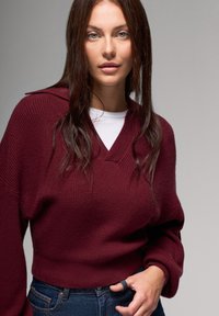 Bordowy sweter z dzianiny z dekoltem w serek i szerokimi rękawami, zestawiony z białą koszulką pod spodem. Dżinsy widoczne na wysokości talii.