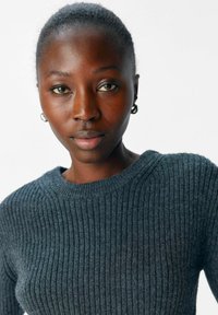 Dunkelgrauer gerippter Strickpullover mit rundem Halsausschnitt, der eine weiche Textur und einen dezenten Glanz aufweist. Das Model hat kurze Haare und schlichte Ohrringe.