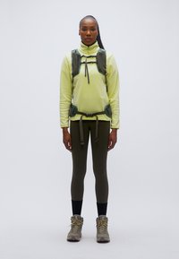 Κίτρινο φλις τοπ με ψηλό γιακά, πράσινη ζώνη από πάνω, σκούρο κολάν και γκρι μποτάκια. Το μοντέλο στέκεται όρθιο μπροστά σε γκρι φόντο.