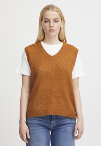Rustorangefarbener Strickpullover mit V-Ausschnitt, der ein strukturiertes Zickzackmuster aufweist, getragen über einem weißen T-Shirt und kombiniert mit blauen Jeans.