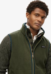 Kamizelka polarowa w ciemnozielonym kolorze z kontrastowym granatowym wykończeniem, zapięcie na zamek błyskawiczny i detale z guzikami. Teksturowane szare rękawy z dzianiny dodają wizualnego zainteresowania.