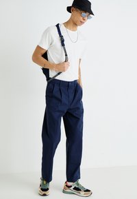 Navy pleated trousers, white t-shirt, black bucket hat, silver chain, and multi-colored sneakers. Backpack with adjustable strap.