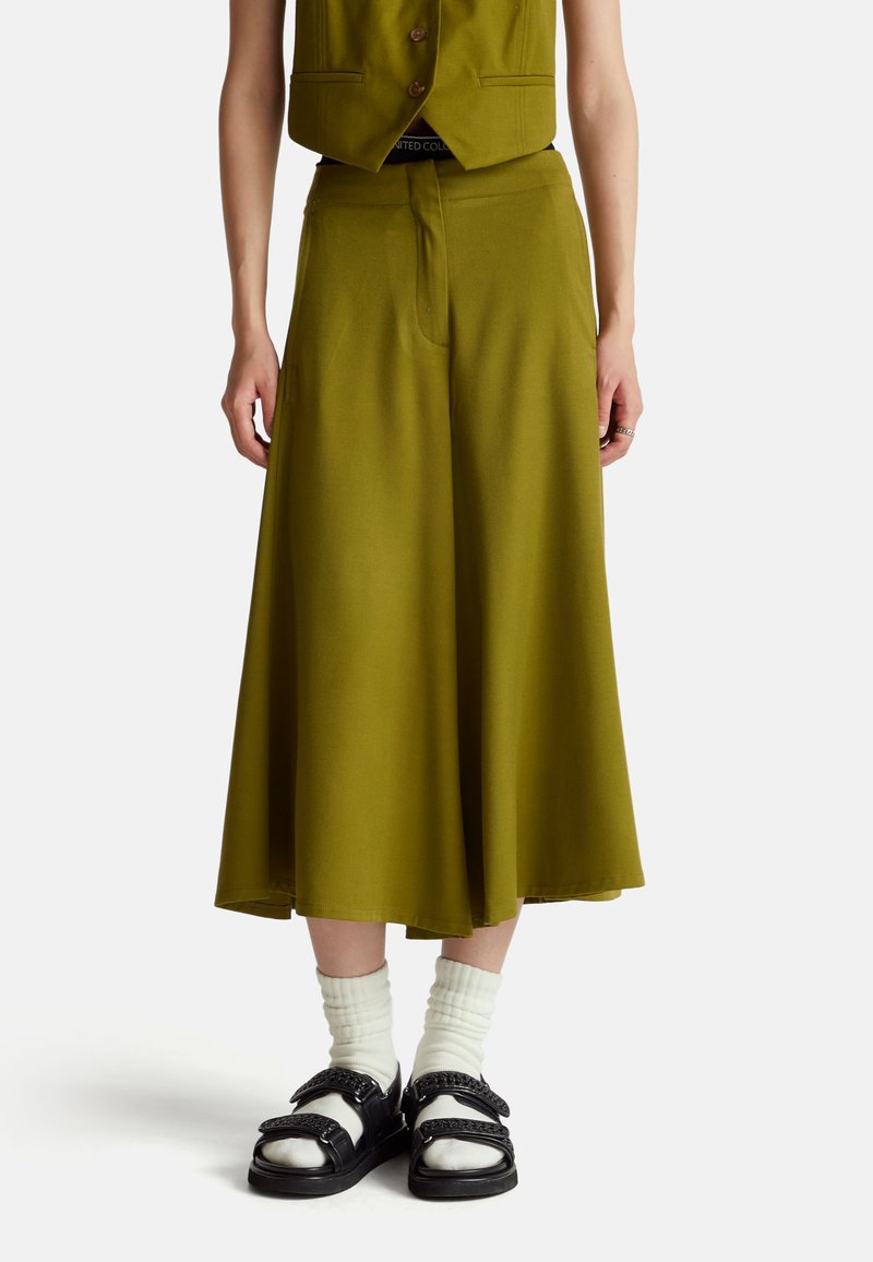 Persoon in olijfgroene wijde cropped broek, met witte sokken en zwarte sandalen, staande tegen een effen witte achtergrond.