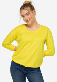 Langärmliges gelbes T-Shirt aus weichem Stoff, mit rundem Halsausschnitt und lockerer Passform, kombiniert mit dunkelblauen Jeans.